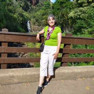 A woman in a green shirt and white pants stands on a bridge in front of a waterfall, surrounded by lush greenery.