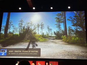 A presentation screen displays a digital nature scene labeled "2022 - Digital Twins of Nature" by Dr. Maria C.R. Harrington & Chris Jones. Five people are seated at a panel below.