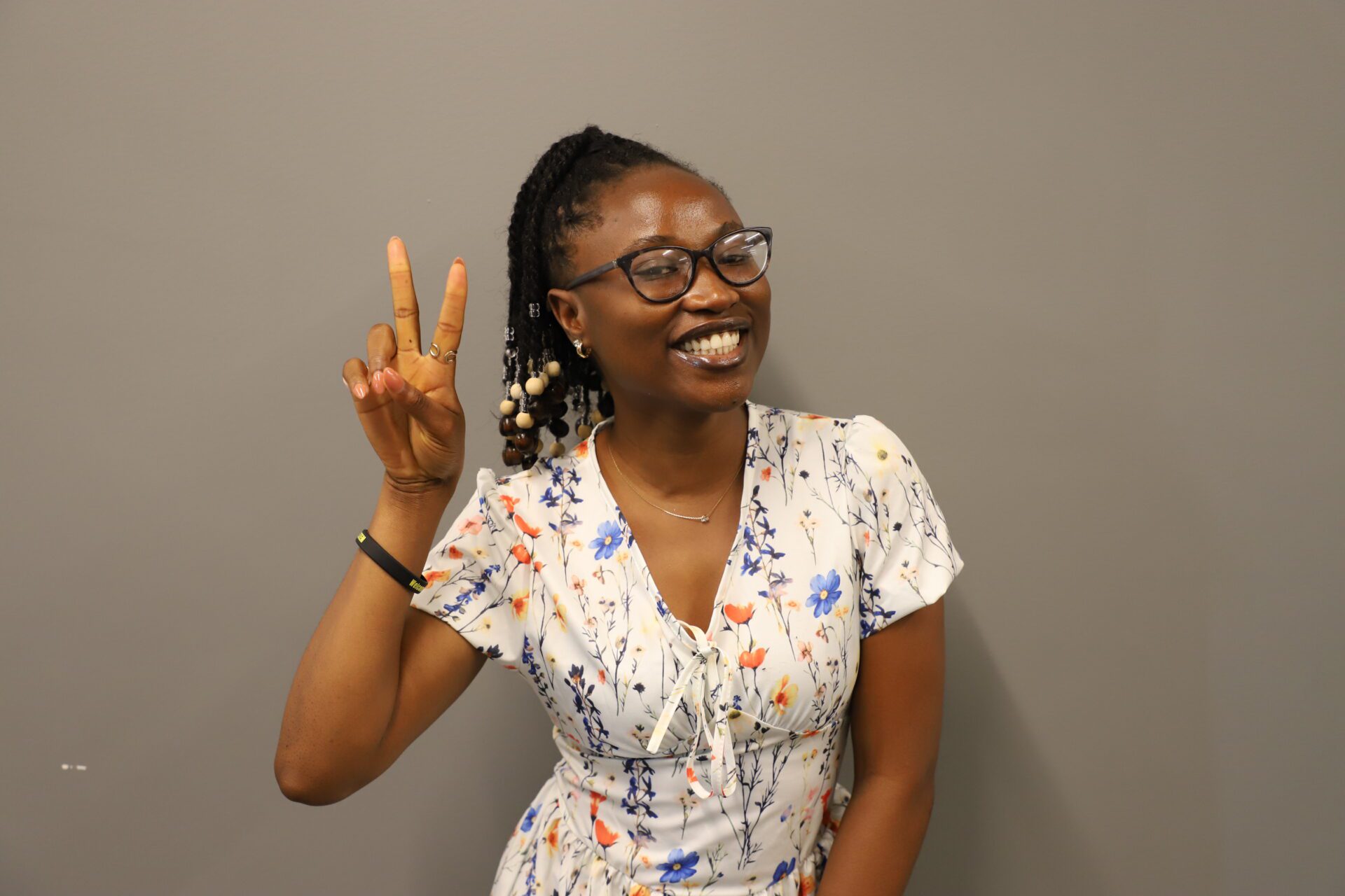 Smiling woman in glasses and a floral dress stands against a gray wall, holding up her hand and making a peace sign gesture.