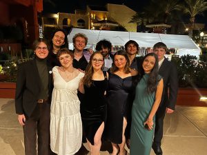 A group of ten people dressed in formal attire posing for a photo outdoors at night, with a white tent and palm trees in the background.