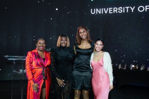 Four people are standing in front of a dark backdrop with "University of" visible. They are dressed in formal attire and smiling.