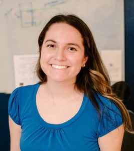 A person with long hair wearing a blue shirt smiles in front of a whiteboard with drawings and papers.