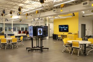 A modern tech lab features yellow chairs, round tables, digital screens, and overhead equipment in a spacious, brightly lit room.