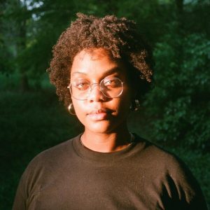 A person with curly hair, glasses, and a black shirt stands outside in a sunlit area with green foliage in the background.