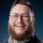 Person with a beard and glasses smiling against a dark blue background.