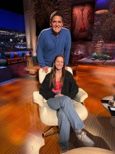 A man stands behind a woman sitting in a chair on a TV set with warm lighting and cityscape background.