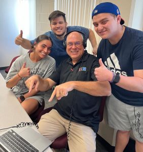 Four people smile and give thumbs up around a seated man wearing headphones at a desk.