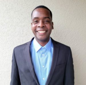 Smiling person wearing a blue shirt and dark suit jacket stands against a light-colored wall.