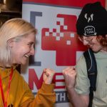 Two people smiling and pretending to fist bump against a gaming-themed backdrop indoors.