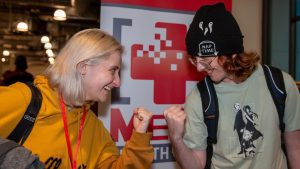 Two people smiling and pretending to fist bump against a gaming-themed backdrop indoors.