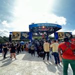 A crowd gathers at an outdoor event for "Fox Big Noon Kickoff," featuring UCF and another team's logos on a stage, with bright daylight and scattered clouds above.