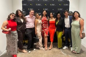 A group of nine people posing in front of a "College of Business" backdrop. Some are wearing formal attire, and two hold black and gold balloons.