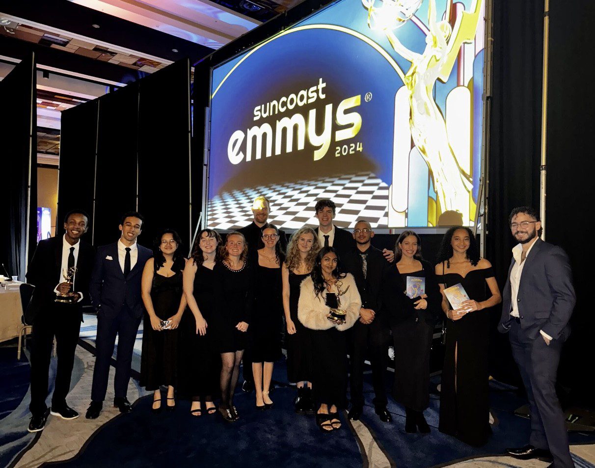 A group of people in formal attire stand in front of a "Suncoast Emmys 2024" display at an event.