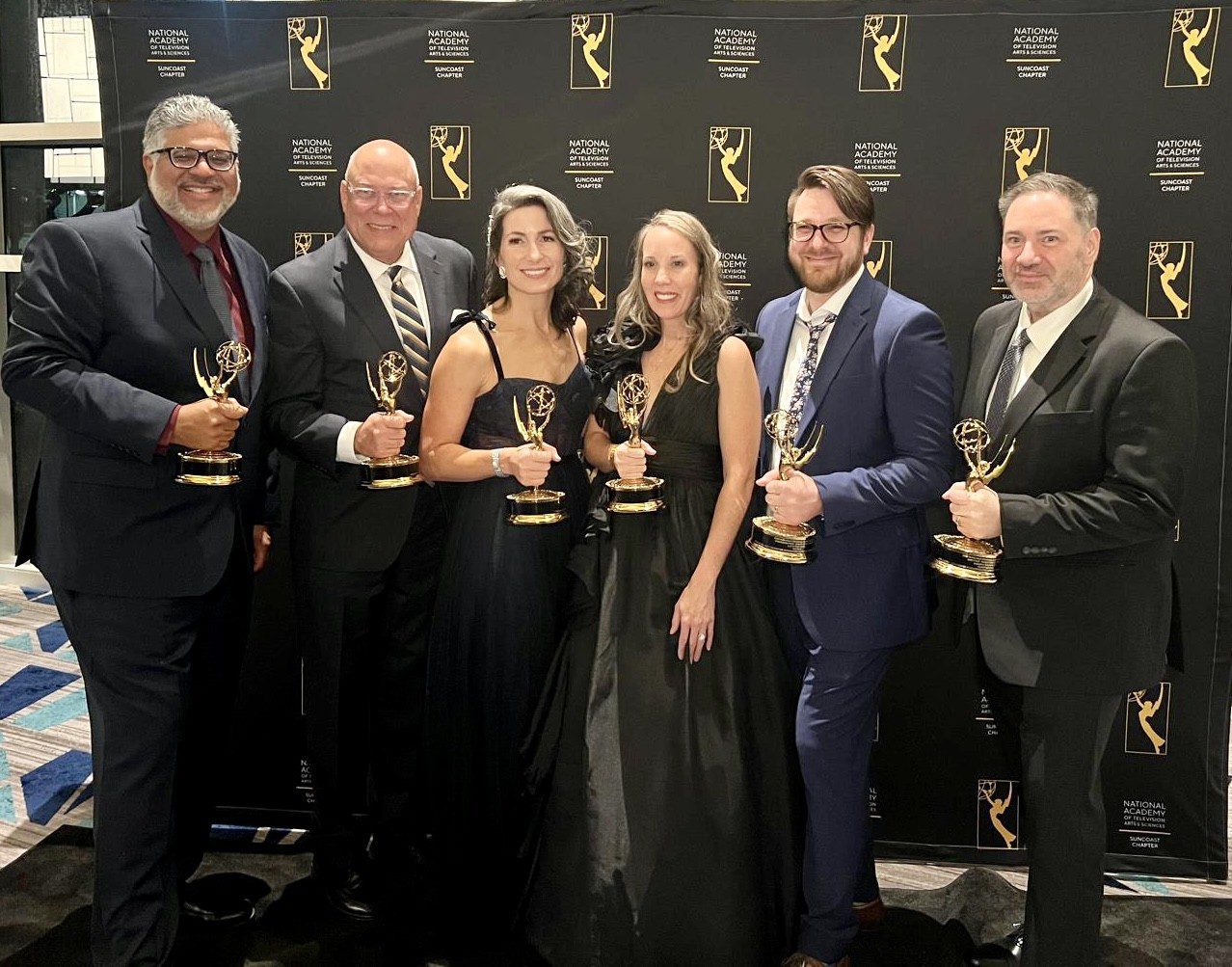 Six people in formal attire stand in front of a backdrop, each holding an Emmy award.