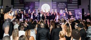 A group of people on stage holding champagne bottles and sparklers, with an audience taking photos. Banners in the background indicate a celebratory event.