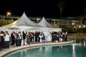 People in formal attire gather at an outdoor event near a pool, with white tents and a banner in the background.