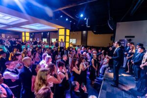 A man in a suit speaks to a large, well-dressed crowd at an indoor event. People are standing and taking photos.