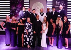 A group of 15 people dressed formally stands together, posing for a group photo in front of a backdrop at an event.