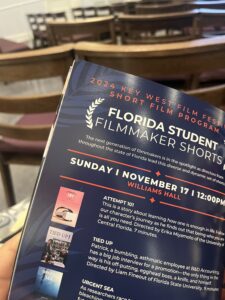 A person holds a film program booklet for the 2024 Key West Film Festival. It lists two films: "Attempt 101" and "Tied Up." Background shows rows of empty chairs.