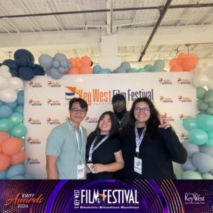 Four people posing together at the Key West Film Festival. They stand in front of a backdrop with balloons. Signs and festival branding are visible. Erica Miyamoto stands center-left.