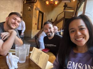 Three people seated at a restaurant table with menus present, smiling at the camera. The setting has rustic decor.