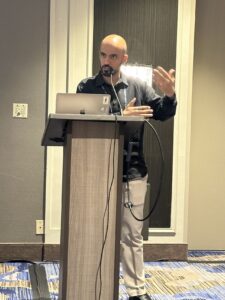 A person is speaking at a podium with a laptop, gesturing with their hands.