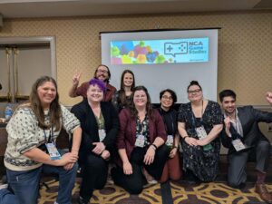 A group of eight people posing together in front of a projector screen displaying "NCA Game Studies" in a conference room.