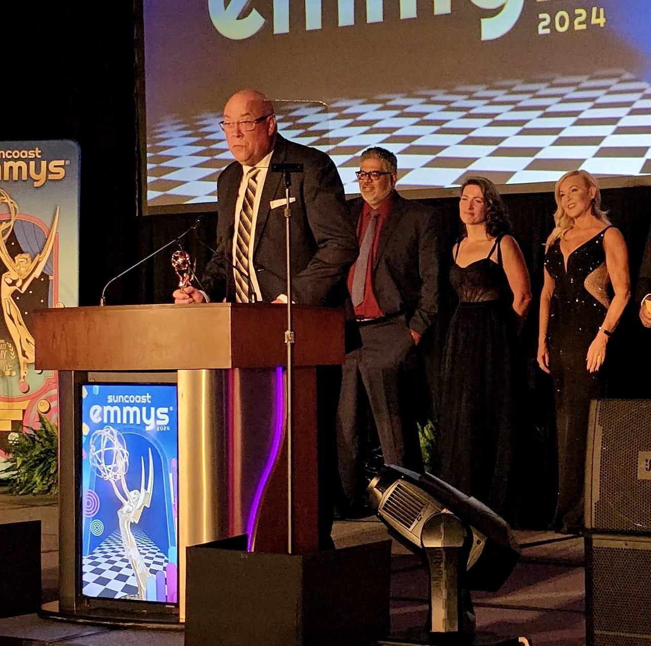 Rick Brunson speaks at a podium holding an award while others stand nearby on stage, with a banner reading "Suncoast Emmys 2024" in the background.