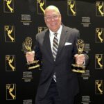 Rick Brunson in a suit holding two Emmy awards, standing in front of a backdrop with National Academy of Television Arts & Sciences Suncoast Chapter logos.