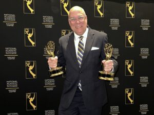 Rick Brunson in a suit holding two Emmy awards, standing in front of a backdrop with National Academy of Television Arts & Sciences Suncoast Chapter logos.