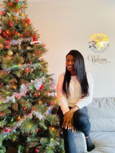 Shalewa Babatayo in a white sweater sits on a couch next to a decorated Christmas tree with colorful lights.