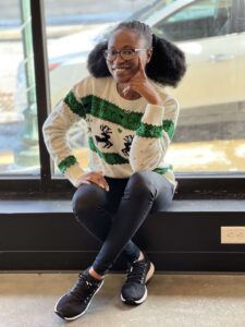 Shalewa Babatayo wearing glasses and a festive sweater with reindeer sits on a bench in front of a window, smiling with one hand on their chin.