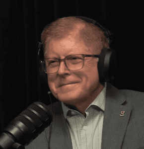 A person wearing glasses and headphones is speaking into a microphone against a dark background.