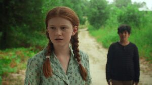 A red-haired girl with braided hair stands on a dirt path, looking off to the side. A boy stands behind her out of focus. Trees and greenery surround them.