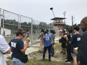 Film crew recording a scene outside a fenced area with barbed wire; crew members wearing "CREW" shirts, using boom mic, and holding equipment near a watchtower.