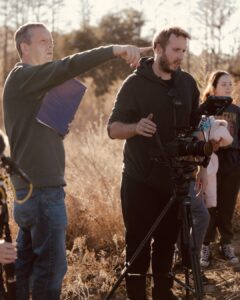 A man points while another operates a camera on a tripod outdoors. Two other people stand nearby. It appears to be a film set.