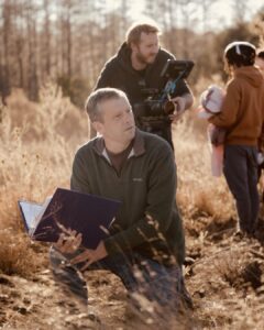 A person crouches with a folder in hand, while another person operates a camera, capturing a scene with a child and a doll in an outdoor setting.