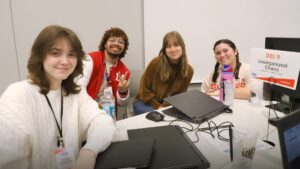 Four people are seated around a table with laptops. A sign reads "Table 19 Unorganized Chaos.