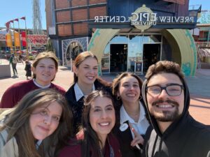 Group of six people posing for a selfie outside the Epic Center entrance on a sunny day.