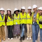 Seven people wearing white hard hats and yellow safety vests stand together, smiling, in front of a beige wall.