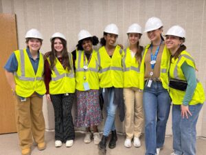 Seven people wearing white hard hats and yellow safety vests stand together, smiling, in front of a beige wall.