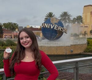 A woman in a red top stands in front of the Universal Studios globe, holding a circular object. Palm trees and theme park structures are visible in the background.