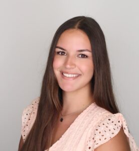 A person with long brown hair smiles while wearing a peach eyelet top against a light gray background.