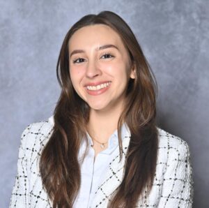 A person with long hair smiles, wearing a white shirt and a checkered blazer, against a gray background.