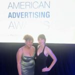 Two people in formal dresses stand in front of a screen displaying "American Advertising Awards" at an event.