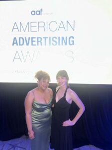 Two people in formal dresses stand in front of a screen displaying "American Advertising Awards" at an event.