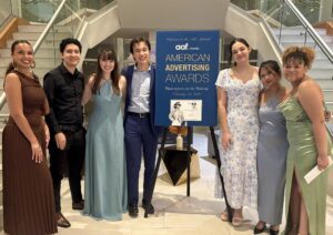 A group of seven people stand in front of a sign for the American Advertising Awards on a stairway. They are dressed in formal attire.