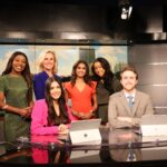 A group of six people, five women and one man, stand and sit around a news desk in a studio setting with cityscape backdrop and studio lighting.