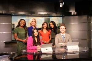 A group of six people, five women and one man, stand and sit around a news desk in a studio setting with cityscape backdrop and studio lighting.