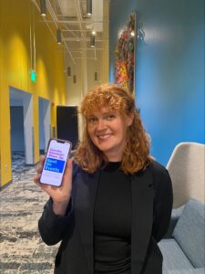 Anna McLean with curly hair smiles at the camera while holding up a smartphone displaying a screen about curating and showcasing events.